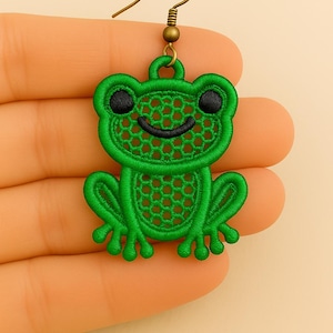 May include: A green frog-shaped earring with black eyes and a smiling mouth. The earring is made of embroidered fabric and has a gold-colored hook. The frog's body has a honeycomb pattern.