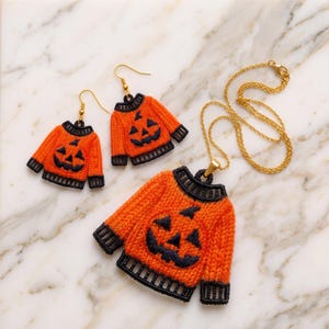 May include: A jewelry set consisting of two drop earrings and a pendant necklace, each featuring a miniature orange sweater with a black jack-o'-lantern face, black ribbed collar, cuffs, and hem, attached to gold-toned hardware and a gold chain.