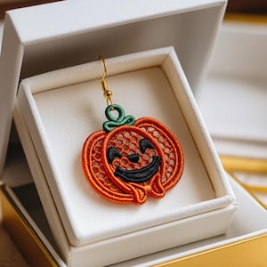 May include: A Halloween-themed earring featuring an orange pumpkin design with a smiling face. The pumpkin is made of orange thread and has a green stem. The earring is displayed in a white gift box with a gold trim.