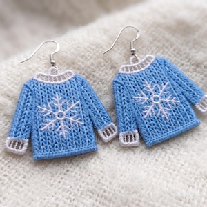 May include: A pair of dangle earrings shaped like miniature blue sweaters with white snowflake patterns on the front. Each sweater features white ribbed detailing at the collar and cuffs, and is attached to a silver-toned metal hook for hanging.