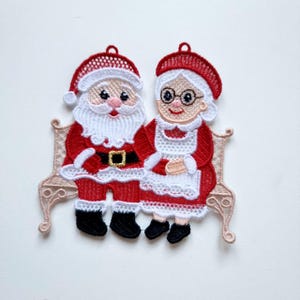 May include: Embroidered Christmas ornament featuring Santa Claus and Mrs. Claus sitting on a decorative bench. Santa wears a red suit with a black belt and boots. Mrs. Claus is in a red dress and white apron. The ornament has a loop for hanging.