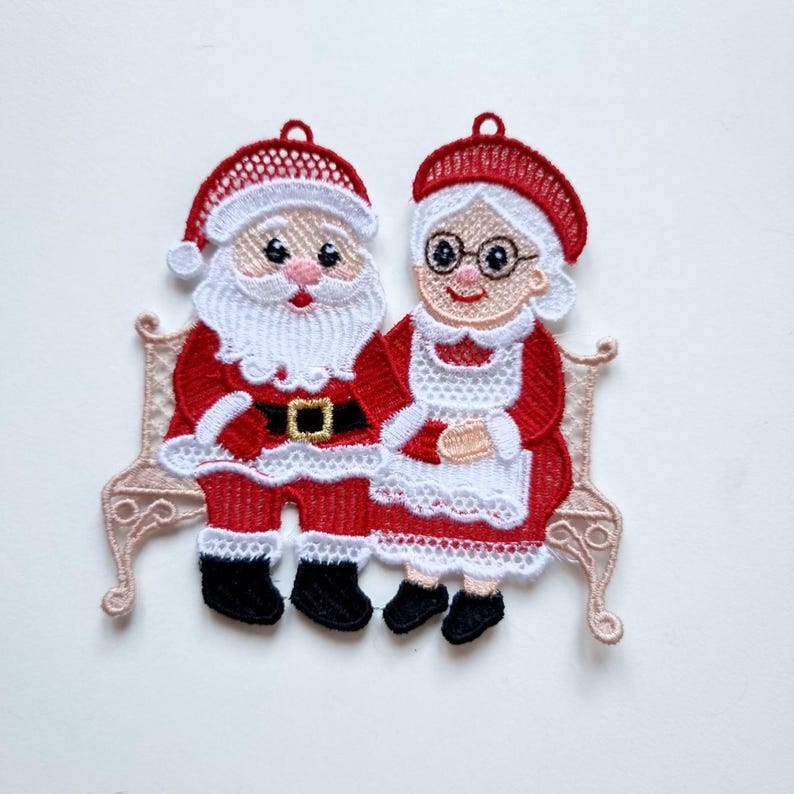 May include: Embroidered Christmas ornament featuring Santa Claus and Mrs. Claus sitting on a decorative bench. Santa wears a red suit with a black belt and boots. Mrs. Claus is in a red dress and white apron. The ornament has a loop for hanging.