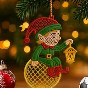 Może przedstawiać: Haftowana ozdoba świąteczna przedstawiająca siedzącego elfa trzymającego latarnię. Elf ubrany jest w czerwono-zielony strój z pasującym kapeluszem i butami. Ozdoba zawieszona jest na złotym sznurku na rozmytym tle światełek choinkowych i gałęzi sosny.