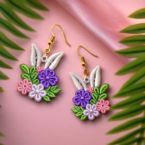 May include: A pair of embroidered earrings featuring a bunny ear design with white, green, purple, and pink floral accents. The earrings have gold-colored hooks and are set against a pink background.