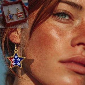 May include: Gold-toned star-shaped earrings with red, white, and blue embroidered details. One earring features a star design with white stars on a blue background, while the other has red and white stripes. The earrings are displayed in a red box.
