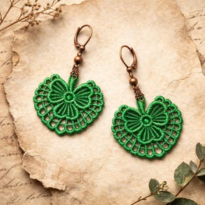 May include: A pair of green macrame earrings. Each earring features a heart-shaped design with a bow in the center and a decorative edge. The earrings have bronze-colored ear wires and accents. The earrings are displayed on a textured, aged paper background.
