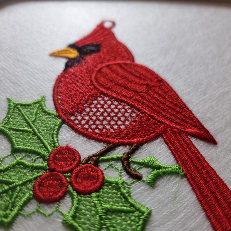 Peut inclure: Broderie d'un cardinal rouge avec un masque noir et un bec jaune, perch&eacute; sur une branche de houx verte avec des baies rouges. L'aile de l'oiseau pr&eacute;sente un motif ajour&eacute; d&eacute;taill&eacute;. La broderie est sur fond blanc.