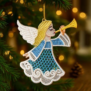 May include: Embroidered Christmas ornament of an angel playing a golden trumpet. The angel has blonde hair, white wings, and a blue and white dress. The ornament is hanging from a gold string against a blurred background of Christmas lights and a pine tree.