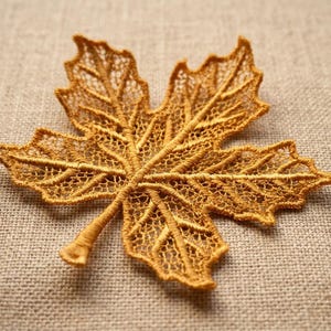 Könnte beinhalten: Eine goldbraune Ahornblatt-Applikation mit aufwendiger, durchbrochener Spitzenstickerei. Das Design zeigt ein detailliertes Netzwerk aus gestickten Blattadern und eine feine, netzartige Struktur über das gesamte Blatt, das in einem Stiel endet.