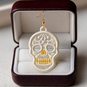 Może przedstawiać: Biały, haftowany kolczyk w kształcie czaszki ze złotymi akcentami, prezentowany w bordowym pudełku na biżuterię. Wzór czaszki ma szczegółowe przeszycia, ze złotą nicią podkreślającą zęby i nos. Kolczyk wisi na złotym haczyku.