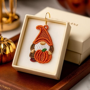 May include: An embroidered gnome earring with an orange hat and pumpkin. The earring is displayed in a cream-colored box. The gnome has a white beard, brown arms, and a gold-colored earring hook.