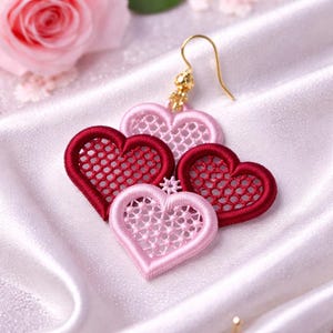 May include: A close-up of a gold earring with four heart-shaped charms. Two charms are a light pink color, and two are a deep red. The hearts have an open, lacy design. The earring is on a white satin surface.