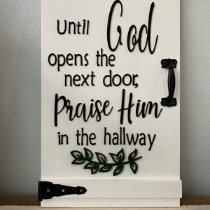 May include: White wooden sign with black text that reads "Until God opens the next door, Praise Him in the hallway". The sign has a black metal handle and a green leafy sprig.