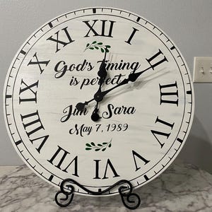 May include: White wooden clock with black Roman numeral markings and black hands. The clock face has the text "God's timing is perfect" and "Jim & Sara May 7, 1989".