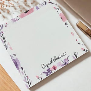May include: Square notepad with a floral border in shades of pink and purple. The name "Raquel Santana" is printed in black script on the bottom right corner. The notepad is on a white surface.