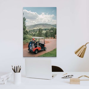 May include: A red golf cart with a black golf bag and other golf equipment in the back. The cart is parked on a paved path with green grass and trees on either side. The background shows a view of a green valley with a mountain range in the distance. The sky is blue with white clouds.