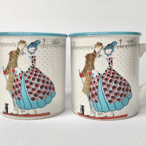 May include: Two white ceramic mugs with blue rims. Each mug features a colourful illustration of a couple in vintage clothing walking with a black cat. The mugs have a white background with a subtle polka dot pattern.