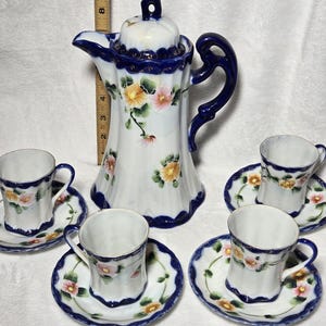 Op de afbeelding: Een witte porseleinen koffiepot en vier bijpassende kopjes en schotels. De set heeft een blauwe rand en een bloemenontwerp met roze en gele bloemen. De koffiepot is ongeveer 20 cm hoog.