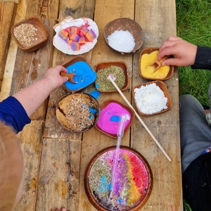 May include: A wooden table with various bowls filled with colorful powders and other materials. The bowls are arranged in a circle, and there are wooden sticks and spoons nearby. The table is made of wood and has a natural finish.