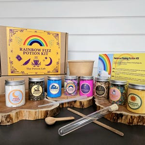 May include: A rainbow fizzing potion kit with a cardboard box, a brown paper cup, and nine small jars filled with colorful ingredients. The jars are labeled with names like "Moon Flakes", "Ancient Meadows", "Ocean Frost", "Rainbow Roots", "Pixie Dust", "Shattered Winds", "Unicorn Foop", and "Sunbeam Sprinkles". The kit also includes a glass test tube, a wooden spoon, and two wooden stirring sticks.