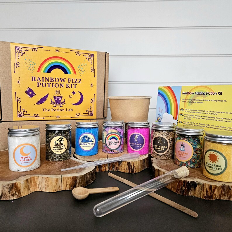 May include: A rainbow fizzing potion kit with a cardboard box, a brown paper cup, and nine small jars filled with colorful ingredients. The jars are labeled with names like "Moon Flakes", "Ancient Meadows", "Ocean Frost", "Rainbow Roots", "Pixie Dust", "Shattered Winds", "Unicorn Foop", and "Sunbeam Sprinkles". The kit also includes a glass test tube, a wooden spoon, and two wooden stirring sticks.