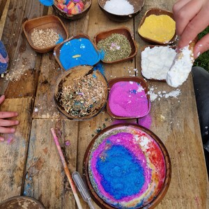 May include: A wooden table with various bowls filled with different colored powders, including blue, pink, white, and yellow. There are also bowls filled with wood chips and dried herbs. A wooden spoon is in the image.