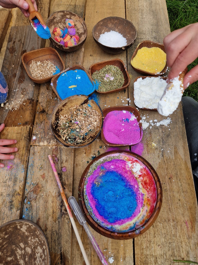 May include: A wooden table with various bowls filled with different colored powders, including blue, pink, white, and yellow. There are also bowls filled with wood chips and dried herbs. A wooden spoon is in the image.
