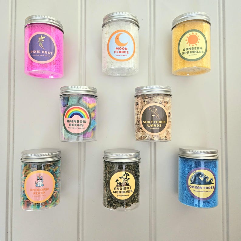 May include: Seven glass jars with silver lids filled with colorful bath salts. Each jar has a label with a whimsical design and name, such as "Pixie Dust", "Moon Flakes", "Sunbeam Sprinkles", "Rainbow Rocks", "Shattered Wands", "Unicorn Poop", "Ancient Meadows", and "Ocean Frost".
