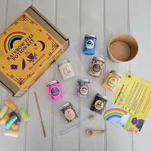 May include: A rainbow fizz potion kit with a brown cardboard box, a wooden spoon, a glass test tube, and seven small glass jars filled with colorful ingredients. The jars are labeled with names like "Ocean Waves", "Rainbow Sprinkles", and "Mystic Moons". The kit also includes a brown paper cup and a yellow instruction sheet with the text "Rainbow Fizz Potion Kit".