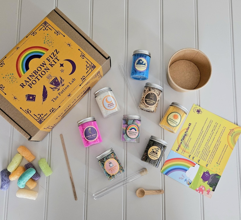 May include: A rainbow fizz potion kit with a brown cardboard box, a wooden spoon, a glass test tube, and seven small glass jars filled with colorful ingredients. The jars are labeled with names like "Ocean Waves", "Rainbow Sprinkles", and "Mystic Moons". The kit also includes a brown paper cup and a yellow instruction sheet with the text "Rainbow Fizz Potion Kit".