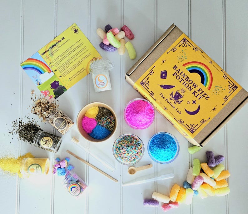 May include: A rainbow fizz potion kit with a yellow box featuring a rainbow and the text "Rainbow Fizz Potion Kit" and "The Potion Lab". The kit includes ingredients in small jars, colorful sprinkles, and other supplies for making fizzy potions.