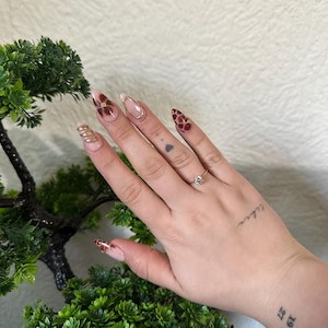 Könnte beinhalten: Eine Hand mit einem goldenen Ring am Ringfinger, die einen Satz mandelförmiger Nägel mit einem burgunderroten und goldenen Blumendesign trägt. Die Nägel sind mit einer hellrosa Basis lackiert.