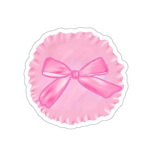 May include: A pink sticker with a watercolour design of a bow. The bow has a glittery outline and is centred on a pink, ruffled circle. The sticker has a white border.