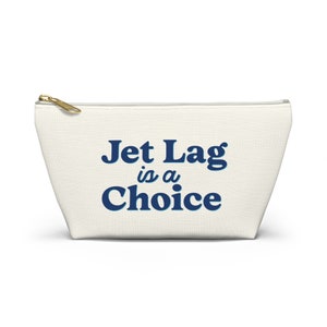 以下が含まれることがあります： 青い文字で「Jet Lag is a Choice」と書かれた白いキャンバス製のジッパー付きポーチ
