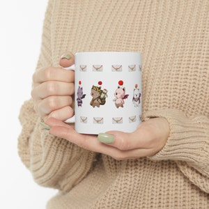 May include: White ceramic mug with a colorful cartoon design of four characters. The characters are wearing hats and have wings. The mug has a white background with a repeating pattern of envelopes.