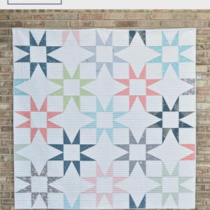 May include: A white quilt with a star pattern in various colors, including blue, pink, and green, hangs against a brick wall. The quilt is labeled "Seeing Stars" and is from Material Girl Quilts by Amanda Castor. The quilt is available in crib, throw, twin, and queen sizes.
