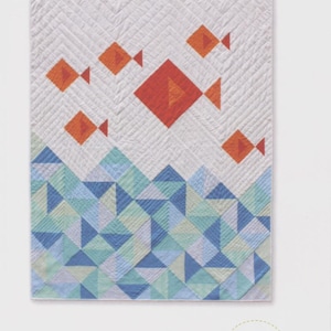 May include: A quilted wall hanging with the text "little fishies quilt." The quilt features orange fish shapes on a white background with a blue and green geometric wave pattern at the bottom. The quilt is from Whole Circle Studio.