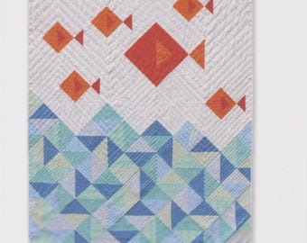 May include: A quilted wall hanging with the text "little fishies quilt." The quilt features orange fish shapes on a white background with a blue and green geometric wave pattern at the bottom. The quilt is from Whole Circle Studio.