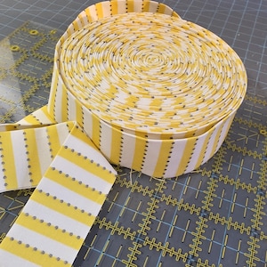 May include: A roll of yellow and white striped fabric, with a dotted pattern, sits on a gray cutting mat. The fabric is unrolled, revealing the pattern. A clear ruler is in the background.