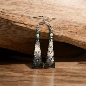 May include: A pair of silver earrings with a geometric pattern and green and white beads. The earrings are triangular in shape and have a textured surface.