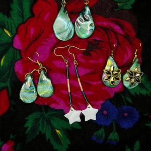 May include: Five pairs of earrings with a variety of styles and colors. The earrings are made of metal, beads, and shell. The earrings are displayed on a floral fabric background.