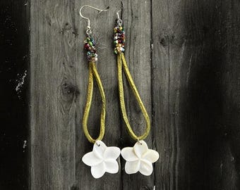 May include: A pair of dangle earrings featuring white flower pendants. The earrings have gold-colored leather straps, beaded accents, and silver-colored hooks. The earrings are displayed against a dark wood background.