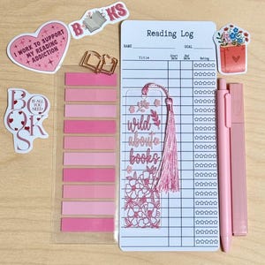 以下が含まれることがあります： 本好きのためのピンクと白の文房具のコレクション。読書記録、ブックマーク、ステッカー、付箋、ペン、蛍光ペンが含まれています。ブックマークには「wild about books」と書かれています。