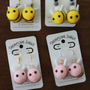 May include: Four pairs of needle-felted animal earrings on white display cards labeled Dreamscape Jungle. Two pairs feature yellow and black bees with white wings, and two pairs feature pink and brown pig-like creatures with white ears and black eyes.