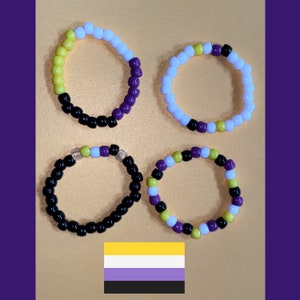 May include: Four beaded rings in the colors of the non-binary pride flag: yellow, white, purple, and black. The rings are arranged on a gold surface with a purple background. A non-binary pride flag is at the bottom.