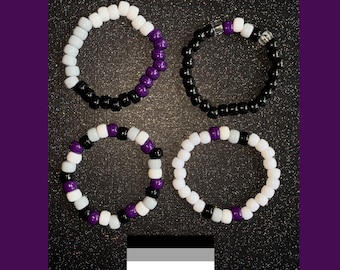 May include: Four black, white, and purple beaded bracelets. The bracelets are arranged in a square pattern on a black background. The bracelets are a symbol of the asexual pride flag.