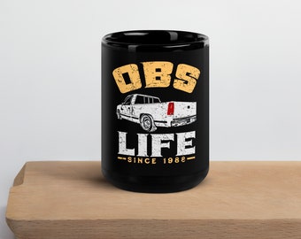 May include: Black coffee mug with a white truck graphic and the text "OBS LIFE SINCE 1982" printed on it. The mug is sitting on a wooden surface.