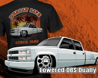 May include: A white, lowered, 1988 Chevrolet pickup truck with a dually rear axle. The truck has a black grille and black wheels. The truck is on a black background with the text "Dually Life Since 1988" and a palm tree sunset graphic.