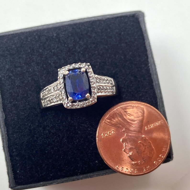 May include: A silver ring with a large, rectangular, deep blue gemstone at the center, surrounded by smaller, clear stones. The ring is set on a black surface next to a penny.