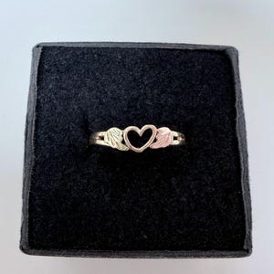May include: A metallic ring featuring a central heart shape flanked by two stylized leaf or wing motifs on either side. The band is polished and smooth, showcasing a warm, multi-toned metal finish that highlights the intricate design elements.